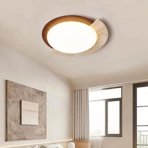 Może przedstawiać: Nowoczesna lampa sufitowa o okrągłym kształcie. Lampa ma jasne, białe, owalne centrum, otoczone brązową i beżową ramą. Oprawa jest zamontowana na białym suficie, z pokojem w neutralnych odcieniach w tle.