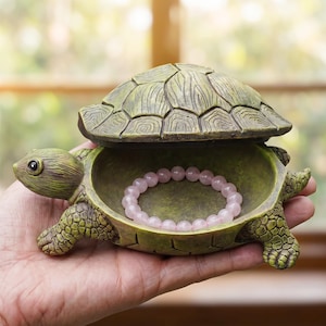May include: A green turtle-shaped trinket box with an open lid, revealing a pink beaded bracelet inside. The box has a textured, aged appearance. The turtle is held in a hand, showcasing the decorative storage container.