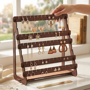 May include: A wooden earring display stand with multiple rows of slots, holding various earrings. The stand is a dark brown color and has an A-frame design. The earrings include dangling, hoop, and stud styles in gold, white, and other colors. The stand is on a white surface.