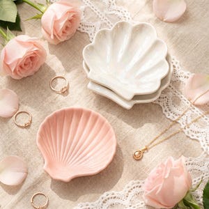 May include: Two ceramic shell-shaped trinket dishes, one in a glossy white finish and one in a soft pastel pink, accompanied by two gold-toned rings and a delicate gold chain necklace with a small pendant, arranged with fresh light pink roses.