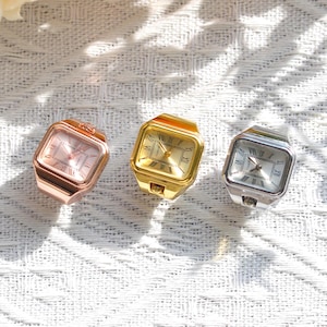May include: Three square ring watches in rose gold, yellow gold, and silver tones. Each watch features a metallic finish, a square face with Roman numeral hour markers, and a central crown. The watch faces display the word Quartz in small text.
