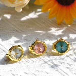 Puede incluir: Tres anillos reloj de tono dorado con esferas redondas y visualización analógica. El reloj de la izquierda tiene esfera blanca, el central rosa y el de la derecha turquesa con un gráfico de trébol. Cada anillo cuenta con una corona.