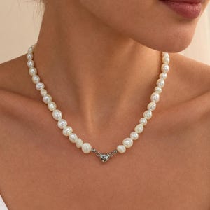 May include: A necklace composed of white, irregularly shaped freshwater pearls strung together. The strand features a small, polished silver-toned heart-shaped charm positioned at the centre, serving as a focal point for the pearl necklace design.