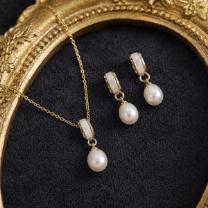 May include: A gold necklace and earring set featuring white accents and pear-shaped pearls. The necklace has a delicate chain and pendant. The earrings dangle from a white and gold setting. The jewellery is presented on a dark, textured surface.