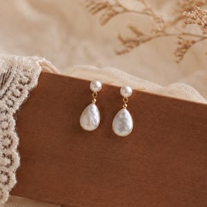 May include: A pair of gold-toned drop earrings featuring a small round pearl stud connected to a larger, teardrop-shaped white pearl pendant. The earrings are suspended from a thin gold metal post, showcasing a classic design with lustrous white pearls.