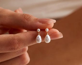 Könnte beinhalten: Ein Paar Ohrhänger bestehend aus einem kleinen runden weißen Perlenstecker, der mit einem größeren tropfenförmigen weißen Perlenanhänger verbunden ist. Die Perlen hängen an dünnen goldfarbenen Metallstiften und bilden ein feines Schmuckstück.