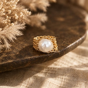 Puede incluir: Un anillo de tono dorado con una perla blanca redonda central, montado sobre una banda formada por varias cadenas metálicas delicadas entrelazadas. El diseño presenta una apariencia texturizada y superpuesta con la superficie lisa y brillante de la perla.