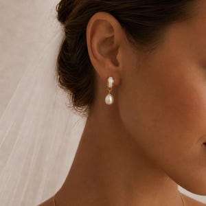 May include: A gold-coloured earring with a white detail and a hanging pearl. The earring is attached to an ear. The background is out of focus.