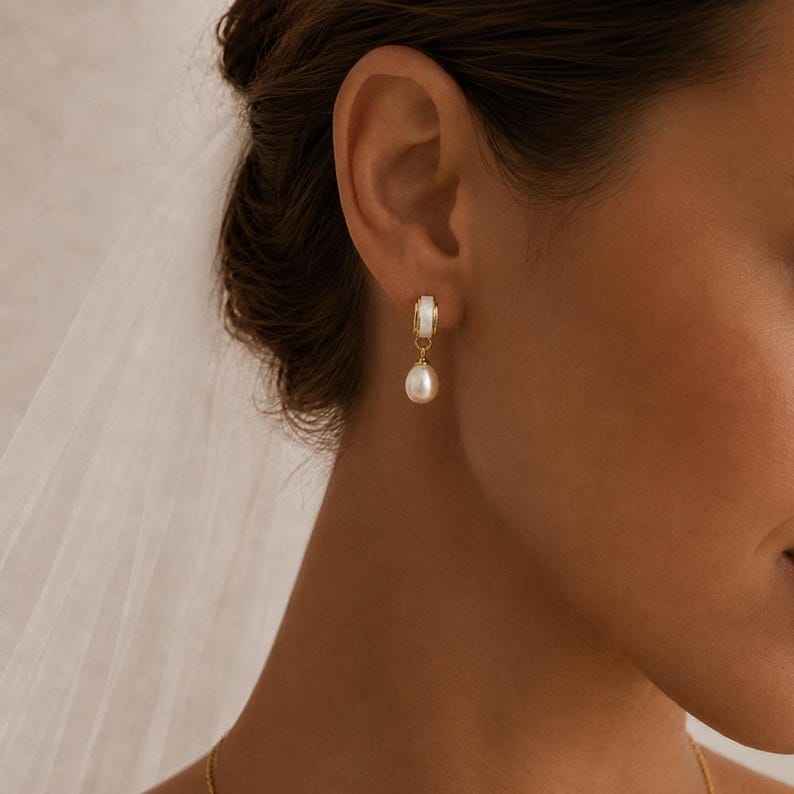 May include: A gold-coloured earring with a white detail and a hanging pearl. The earring is attached to an ear. The background is out of focus.