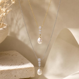 Op de afbeelding: Een gelaagde ketting set bestaande uit twee fijne kettinkjes. De kortere gouden ketting heeft een hanger met drie kleine ronde heldere stenen boven een witte druppelvormige parel. De langere zilveren ketting heeft een identiek hangerontwerp.