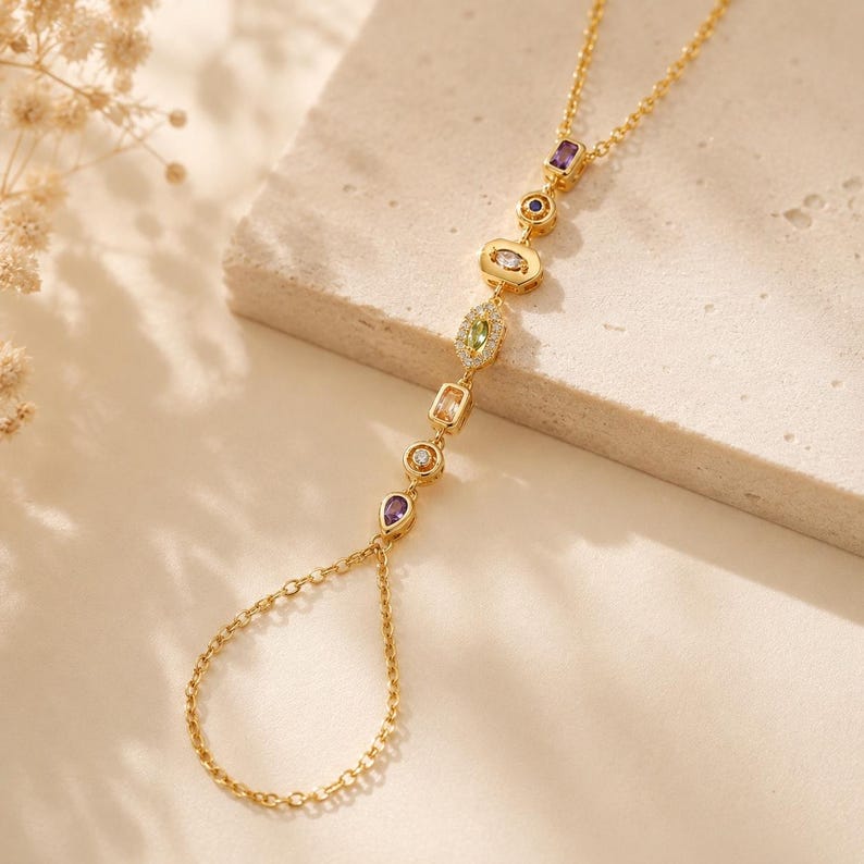 May include: A gold-tone hand chain featuring a delicate link chain design. The piece is adorned with a series of multi-coloured gemstones in various shapes, including pear, round, marquise, and rectangular cuts, set in gold-tone metal settings.