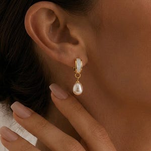 May include: Gold-coloured hoop earring featuring a white enamel detail and a hanging, teardrop-shaped pearl. The earring is displayed on an ear, highlighting the design and elegance of the jewellery. The pearl has a lustrous, off-white hue.