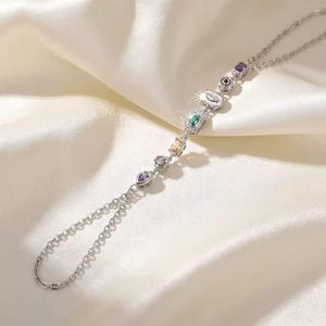 May include: A silver-toned hand chain featuring a delicate link bracelet connected to a finger loop. The chain is adorned with a series of multi-coloured gemstones in various shapes, including pear, round, emerald, and marquise cuts, set in metal.