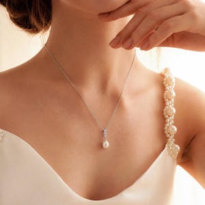 Op de afbeelding: Een zilveren ketting met een fijne schakel en een hanger met drie kleine heldere steentjes verticaal geplaatst boven een enkele druppelvormige witte parel. De ketting wordt gedragen bij een wit kledingstuk met een met parels versierde band.