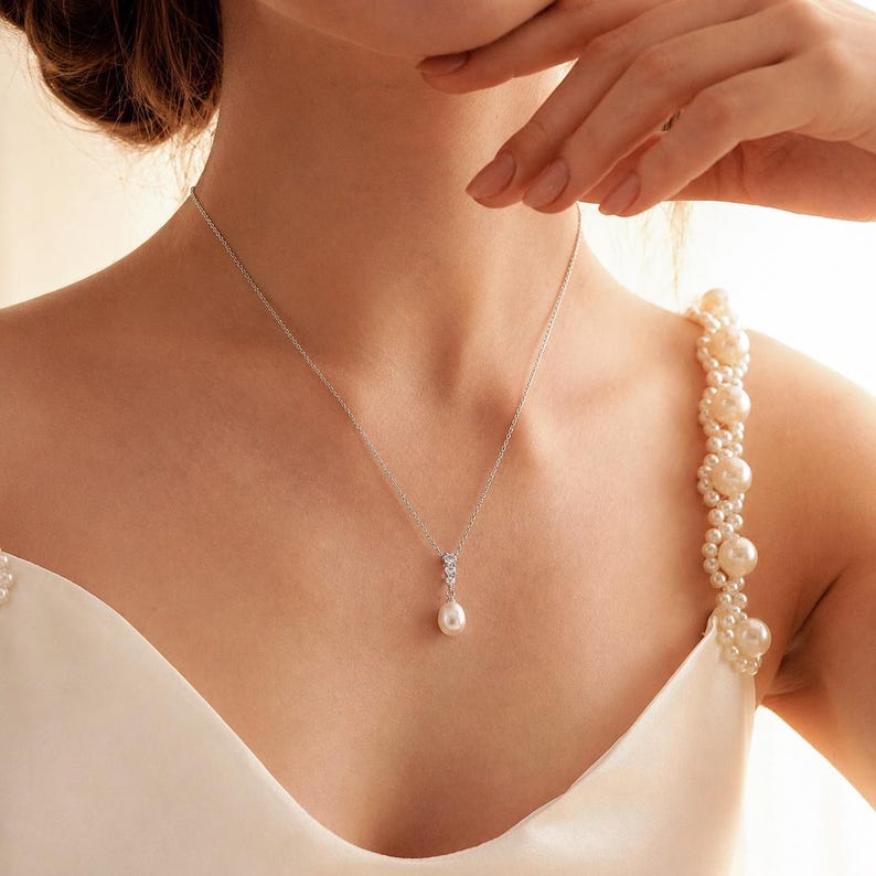 Op de afbeelding: Een zilveren ketting met een fijne schakel en een hanger met drie kleine heldere steentjes verticaal geplaatst boven een enkele druppelvormige witte parel. De ketting wordt gedragen bij een wit kledingstuk met een met parels versierde band.