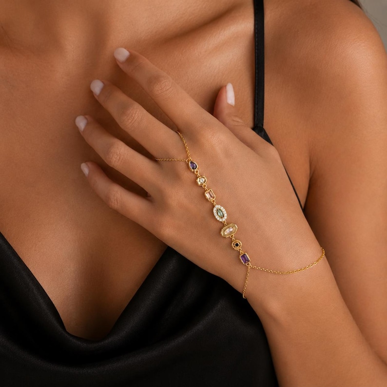 May include: A gold-tone hand chain featuring a delicate chain connecting a ring to a wrist bracelet. The chain is adorned with a series of multi-shaped, multi-coloured gemstones, including purple, clear, and green stones set in gold-coloured metal.