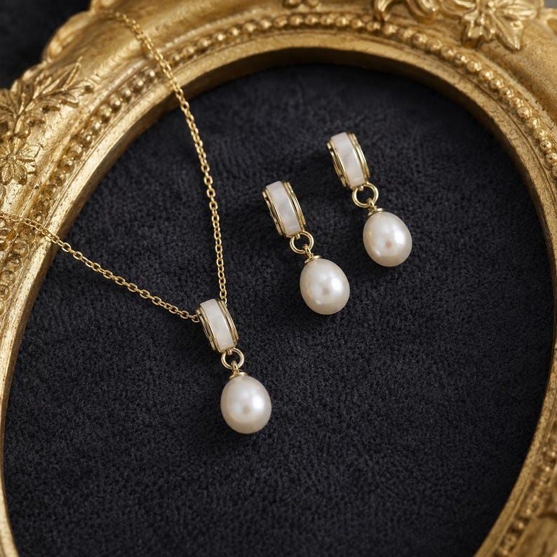May include: A gold necklace and earring set featuring white accents and pear-shaped pearls. The necklace has a delicate chain and pendant. The earrings dangle from a white and gold setting. The jewellery is presented on a dark, textured surface.