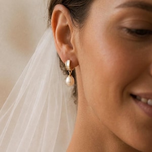 May include: A gold-tone hoop earring with a white section and a hanging, pear-shaped pearl. The earring is worn on an ear, with a white veil in the background. The pearl is a creamy, off-white colour.