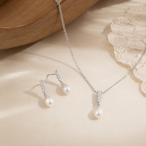 Op de afbeelding: Een zilverkleurige sieradenset bestaande uit een ketting en een paar oorbellen. Elk sieraad is voorzien van een witte druppelvormige parel die hangt aan een verticaal staafje, bezet met drie kleine, heldere, fonkelende zirkonia-steentjes.