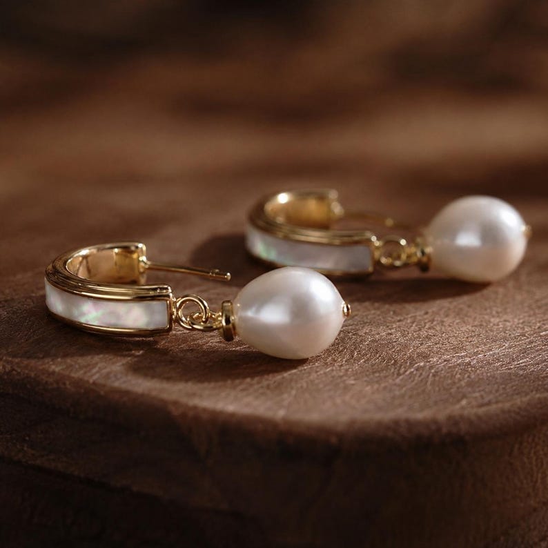 May include: Gold hoop earrings with a mother-of-pearl inlay and a dangling white pearl. The earrings are presented on a brown surface, highlighting their elegant design. These earrings are a classic piece of jewellery. The earrings are a classic jewelry item.