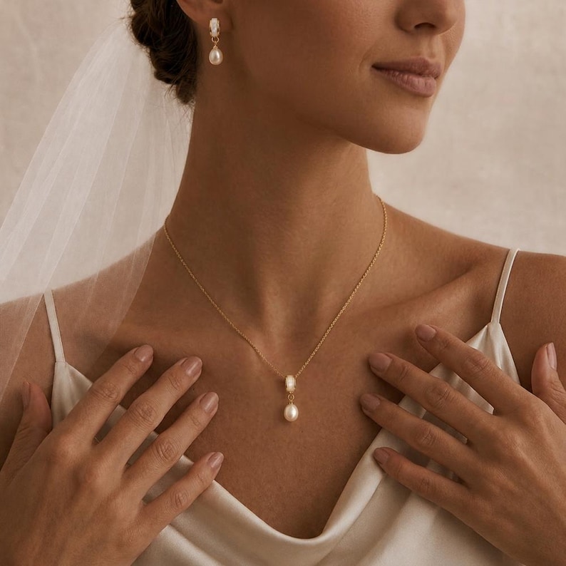 May include: A gold necklace and matching pearl drop earrings. The necklace has a pearl pendant. The jewellery is worn with a white dress. The image is a close-up of the jewellery and the person wearing it.