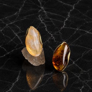 May include: Two gemstone rings with gold-colored settings. One ring features a light yellow stone, the other a dark amber stone. The rings are displayed on a reflective black surface. The text "LumaestudioArt" is at the bottom.