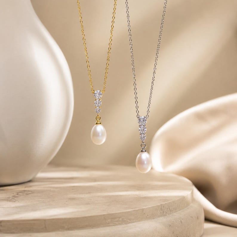 Op de afbeelding: Twee kettingen met een druppelvormige witte parelhangers die aan een verticale rij van drie ronde, heldere stenen hangen. De ene ketting heeft een goudkleurige ketting en zetting, de andere een zilverkleurige ketting en zetting.