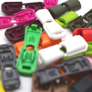 May include: A selection of brightly coloured plastic cord locks in shades such as pink, green, white, and black. These small, rectangular locks have a slotted design and are scattered on a white surface.