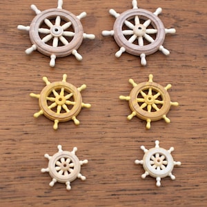 May include: Six wooden ship wheel decorations arranged in three pairs of varying sizes. The top pair is light brown, the middle pair is yellow, and the bottom pair is off-white. Each wheel features a central hub with eight spokes and handles.