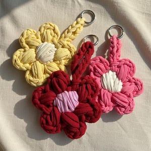 Puede incluir: Tres llaveros con forma de flor hechos de cordón de tela trenzada. Uno es amarillo con centro blanco, otro es rojo oscuro con centro rosa claro y otro es rosa brillante con centro blanco. Cada uno incluye una anilla de metal plateado.