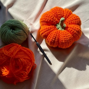 May include: A crocheted orange pumpkin with a light green stem, positioned next to a black crochet hook, a ball of bright orange yarn, and a ball of olive green yarn. The items are arranged together, showcasing the materials used for crafting.