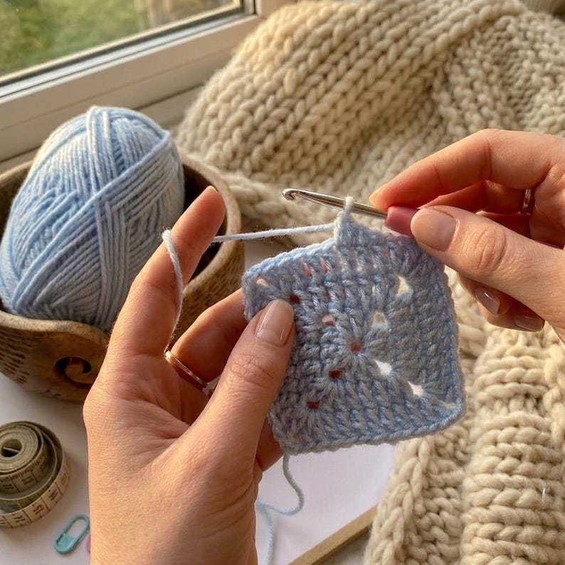Peut inclure: Un carr&eacute; crochet&eacute; bleu clair est en cours de r&eacute;alisation avec un crochet. Une pelote de laine bleu clair est pos&eacute;e dans un bol en bois. Un m&egrave;tre ruban et une &eacute;pingle de s&ucirc;ret&eacute; sont &eacute;galement visibles. L'arri&egrave;re-plan comprend un article tricot&eacute; beige.