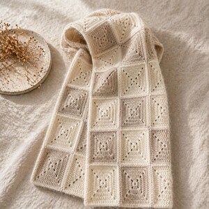 Peut inclure: Une &eacute;charpe en crochet cr&egrave;me et taupe avec un motif g&eacute;om&eacute;trique carr&eacute;. L'&eacute;charpe est pli&eacute;e, r&eacute;v&eacute;lant le design ajour&eacute; complexe. La palette de couleurs neutres et l'aspect textur&eacute; sugg&egrave;rent un accessoire fait main et confortable.