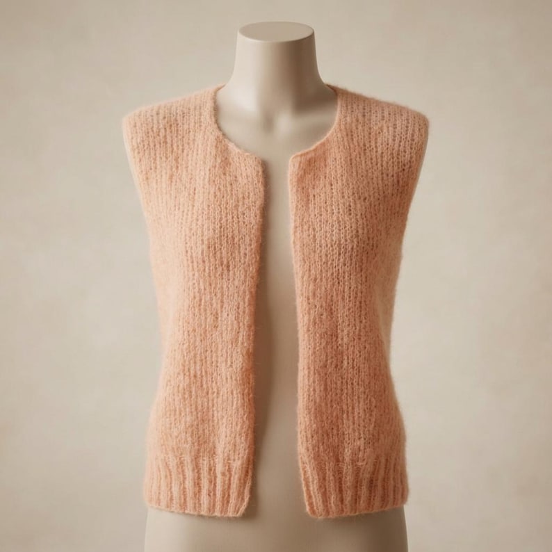 May include: A peach-colored, open-front vest made of a soft, textured knit fabric. The sleeveless garment has a rounded neckline and a straight hemline. The vest is displayed on a mannequin, showcasing its simple, elegant design. The overall style is casual and comfortable.