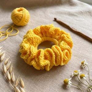 May include: A bright yellow, handmade crochet scrunchie. The scrunchie is circular with a ruffled design. A ball of yellow yarn, a crochet hook, and sprigs of dried flowers are also in the image.