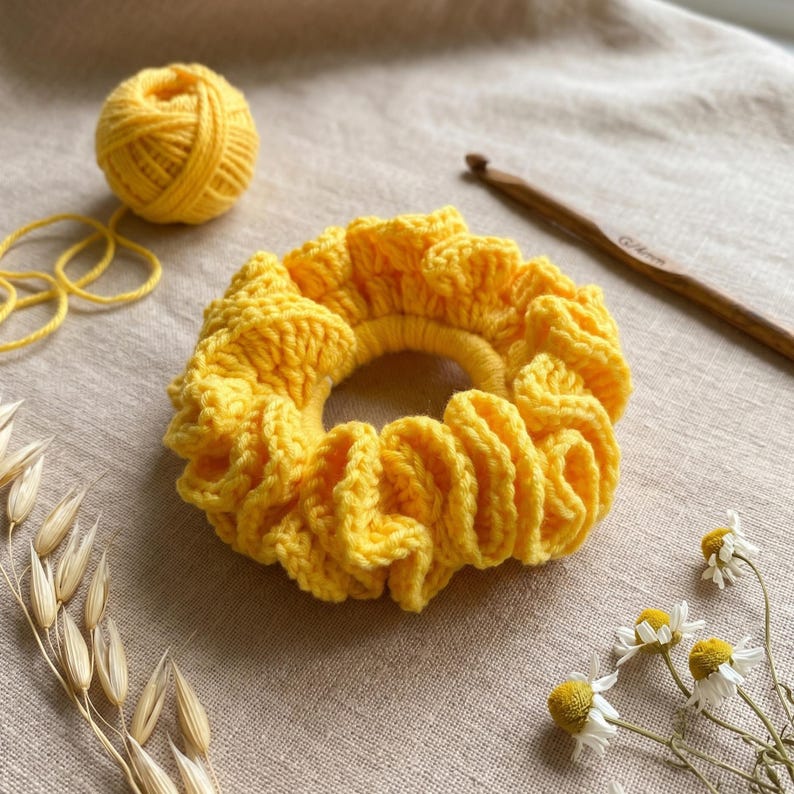 May include: A bright yellow, handmade crochet scrunchie. The scrunchie is circular with a ruffled design. A ball of yellow yarn, a crochet hook, and sprigs of dried flowers are also in the image.