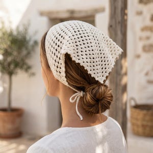 May include: A cream-colored, open-weave crochet headscarf tied at the nape of the neck. The head covering is square-shaped and has a delicate, airy design. The headscarf is worn over brown hair and paired with a white top.