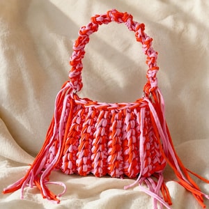 May include: A handmade handbag crafted from thick, braided fabric strips in alternating shades of bright orange and light pink. The bag features a rounded handle and long, dangling fringe accents on both sides, creating a textured appearance.