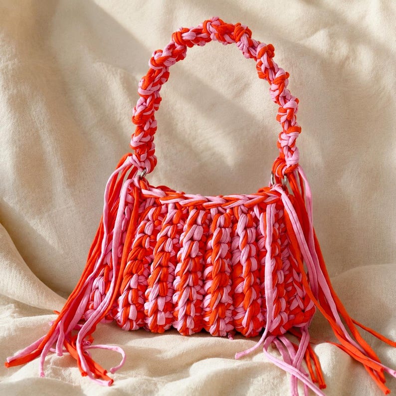May include: A handmade handbag crafted from thick, braided fabric strips in alternating shades of bright orange and light pink. The bag features a rounded handle and long, dangling fringe accents on both sides, creating a textured appearance.