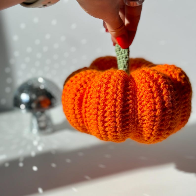 May include: A bright orange crocheted pumpkin featuring a textured, ribbed surface and a small, light green crocheted stem at the top. The pumpkin is round and plump, showcasing detailed yarn stitching throughout its entire exterior structure.
