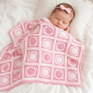 Op de afbeelding: Een baby gewikkeld in een handgemaakte roze en witte gehaakte deken. De deken heeft een vierkant patroon met cirkelvormige ontwerpen. De baby draagt een roze strik hoofdband en rust op een wit kussen.