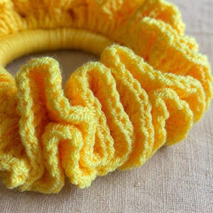 May include: A close-up of a bright yellow crochet scrunchie. The scrunchie is made of soft, textured yarn and is gathered around a yellow elastic band. The crochet stitch creates a ruffled effect.