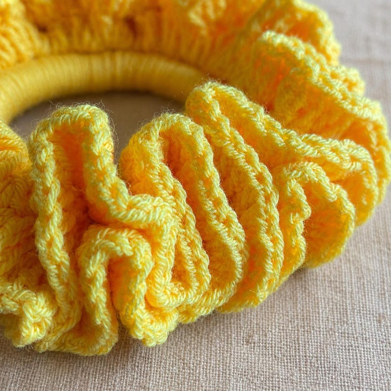 May include: A close-up of a bright yellow crochet scrunchie. The scrunchie is made of soft, textured yarn and is gathered around a yellow elastic band. The crochet stitch creates a ruffled effect.