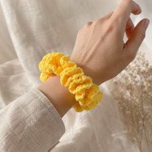 May include: A bright yellow, crocheted scrunchie is worn on a wrist. The scrunchie is made of a textured yarn and has a ruffled appearance. The accessory is a hair tie, perfect for adding a pop of color to any hairstyle.