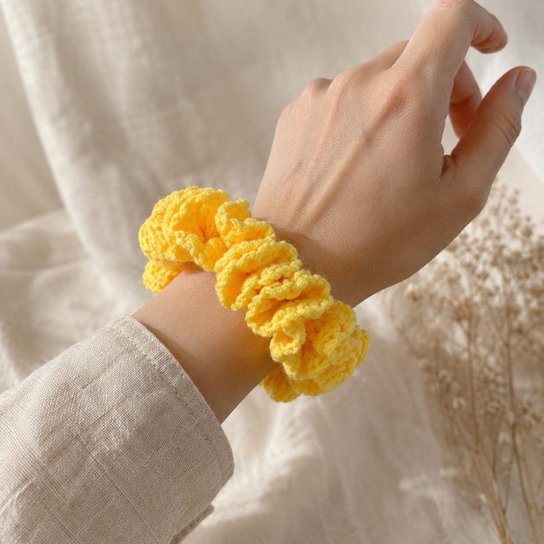 May include: A bright yellow, crocheted scrunchie is worn on a wrist. The scrunchie is made of a textured yarn and has a ruffled appearance. The accessory is a hair tie, perfect for adding a pop of color to any hairstyle.