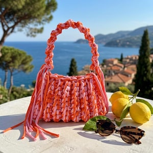 May include: A handmade crochet handbag crafted from thick, braided fabric yarn in alternating shades of bright orange and soft pink. The bag features a rounded handle, a pleated body, and long, dangling fringe tassels on both sides of the opening.