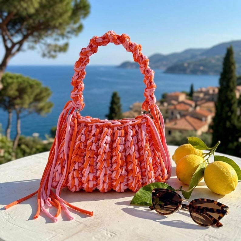 May include: A handmade crochet handbag crafted from thick, braided fabric yarn in alternating shades of bright orange and soft pink. The bag features a rounded handle, a pleated body, and long, dangling fringe tassels on both sides of the opening.