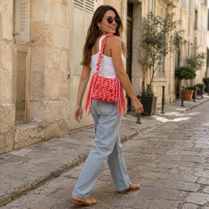 May include: A white tank top, light blue denim jeans, and brown sandals. A bright pink and orange crocheted shoulder bag with long fringe accents hangs from the shoulder. The bag features a thick, braided strap and a textured, multi-colored yarn design.