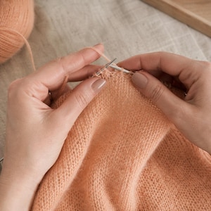 May include: Close-up of hands knitting with peach-colored yarn and silver needles. A ball of yarn is in the upper left corner. The knitted fabric is a light peach color. The background is a neutral beige.