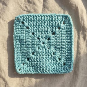 Peut inclure: Un carr&eacute; granny crochet bleu clair. Le carr&eacute; fait main pr&eacute;sente un motif de point textur&eacute; avec des coins ajour&eacute;s. Le carr&eacute; est pr&eacute;sent&eacute; sur un fond de tissu de couleur neutre, mettant en valeur la texture et la couleur du fil.